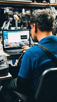 Technician in blue shirt working on a laptop in a busy workshop setting.