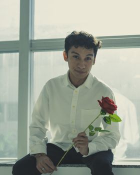 A young man sits by a window, holding a red rose, exuding a contemplative mood.