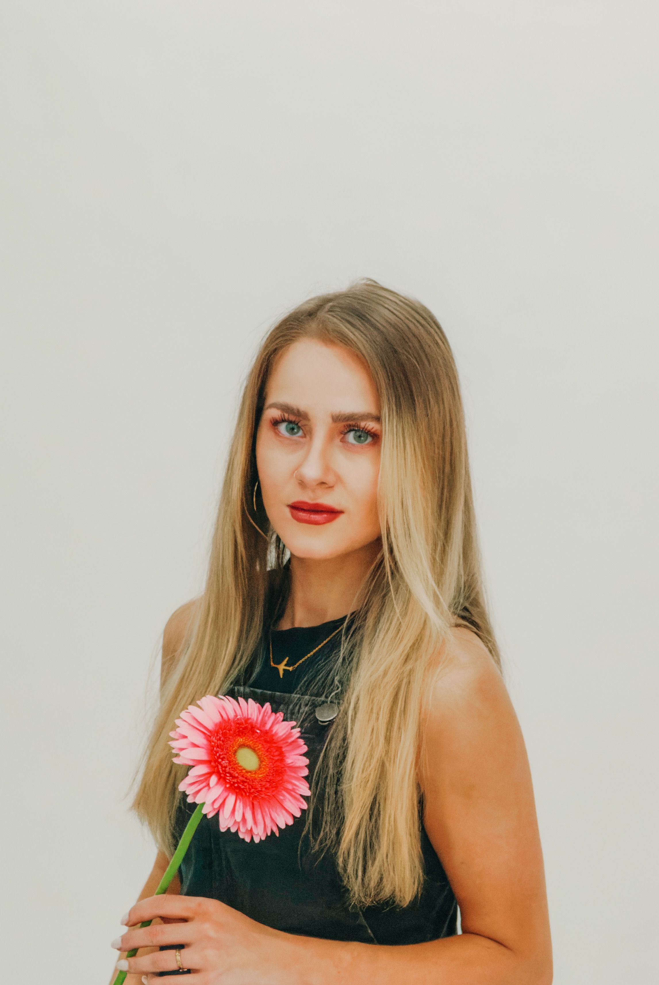 Portrait of a young woman with long hair holding a pink gerbera daisy against a neutral background.
