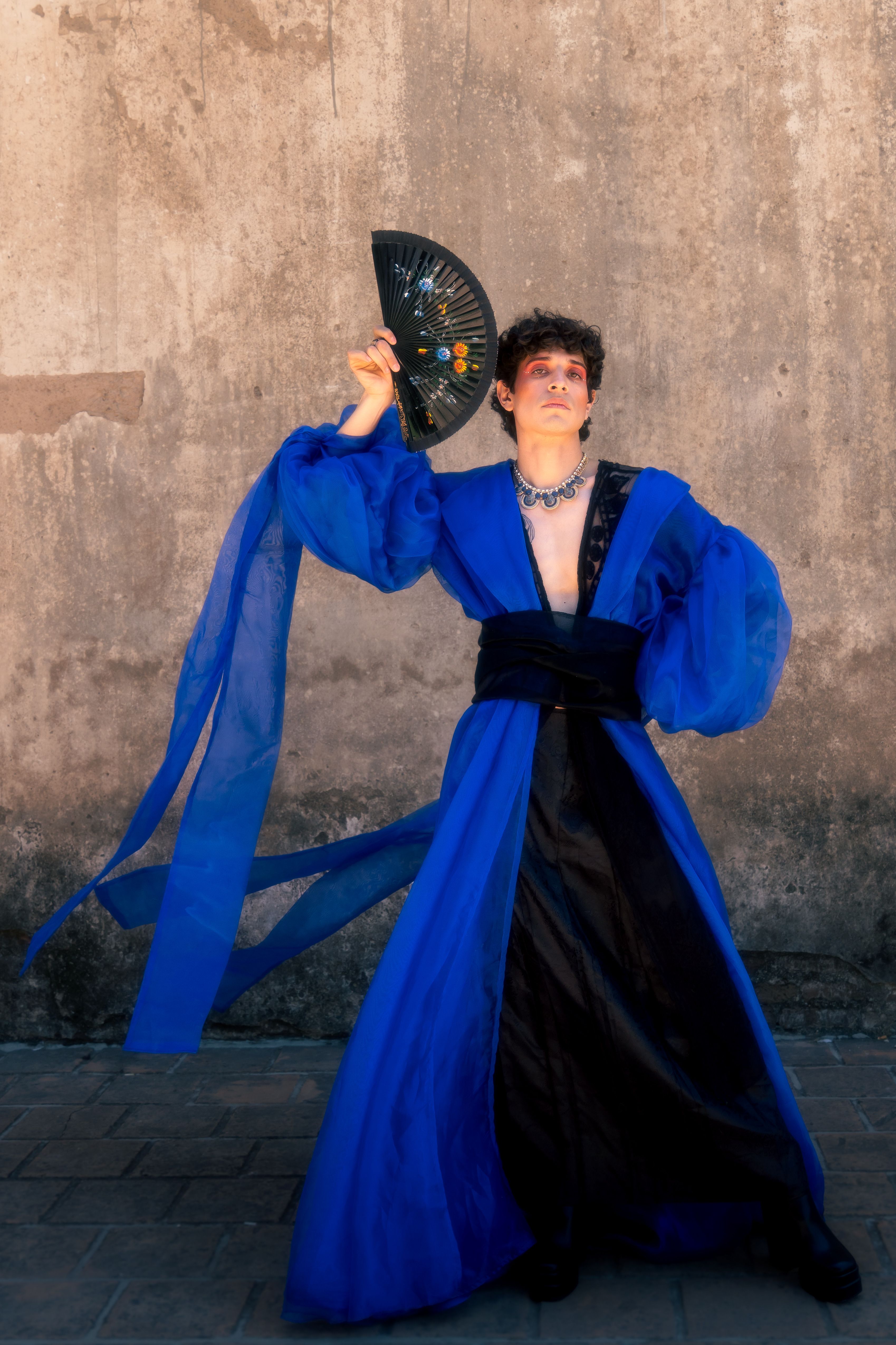 Striking fashion editorial featuring a model in a dramatic blue outfit, posing with a decorative fan.
