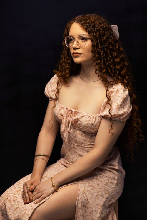 Free A young woman with curly hair wearing a floral dress, sitting gracefully against a dark background. Stock Photo