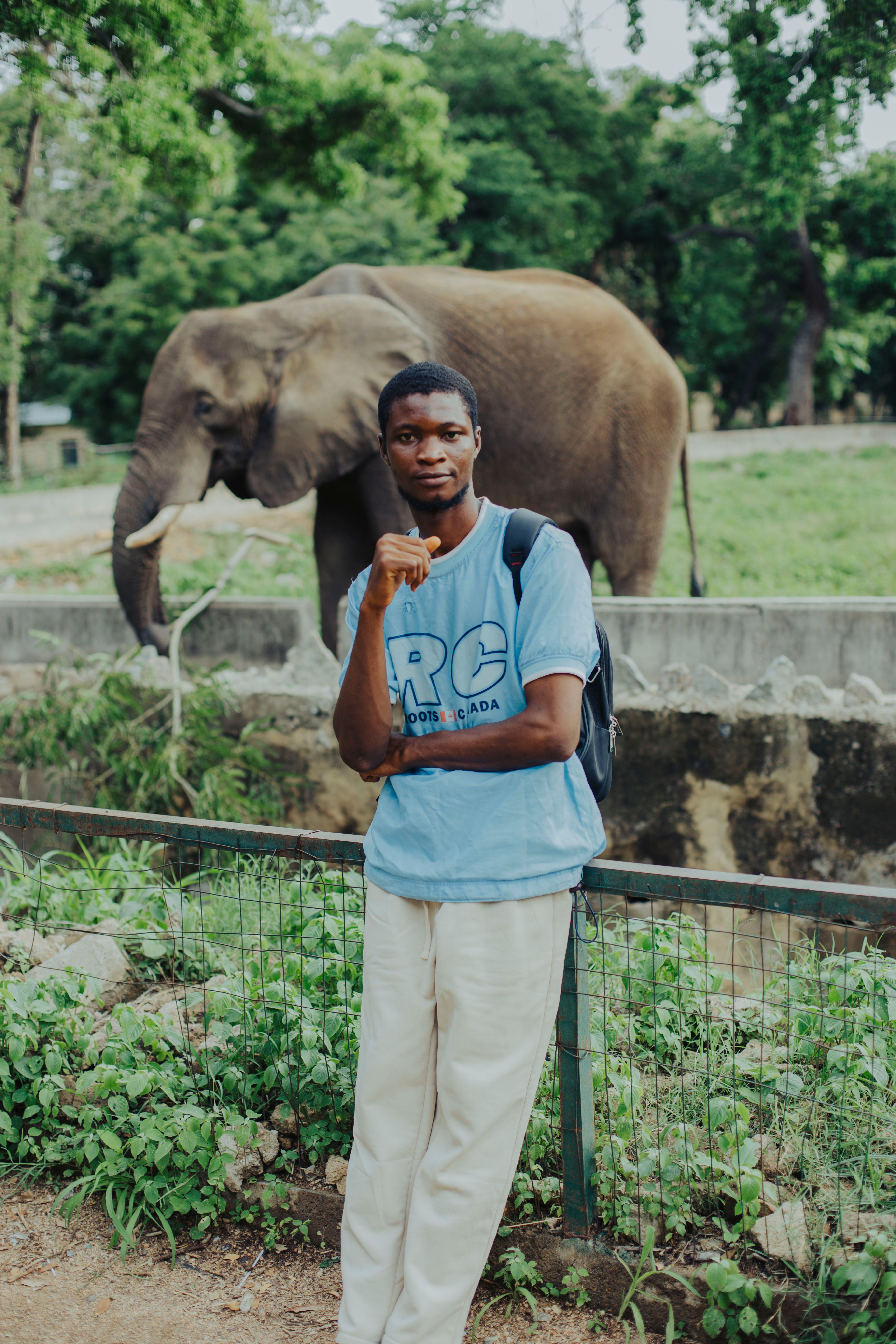 Man poses in front of an elephant at a zoo, showcasing a serene outdoor environment.