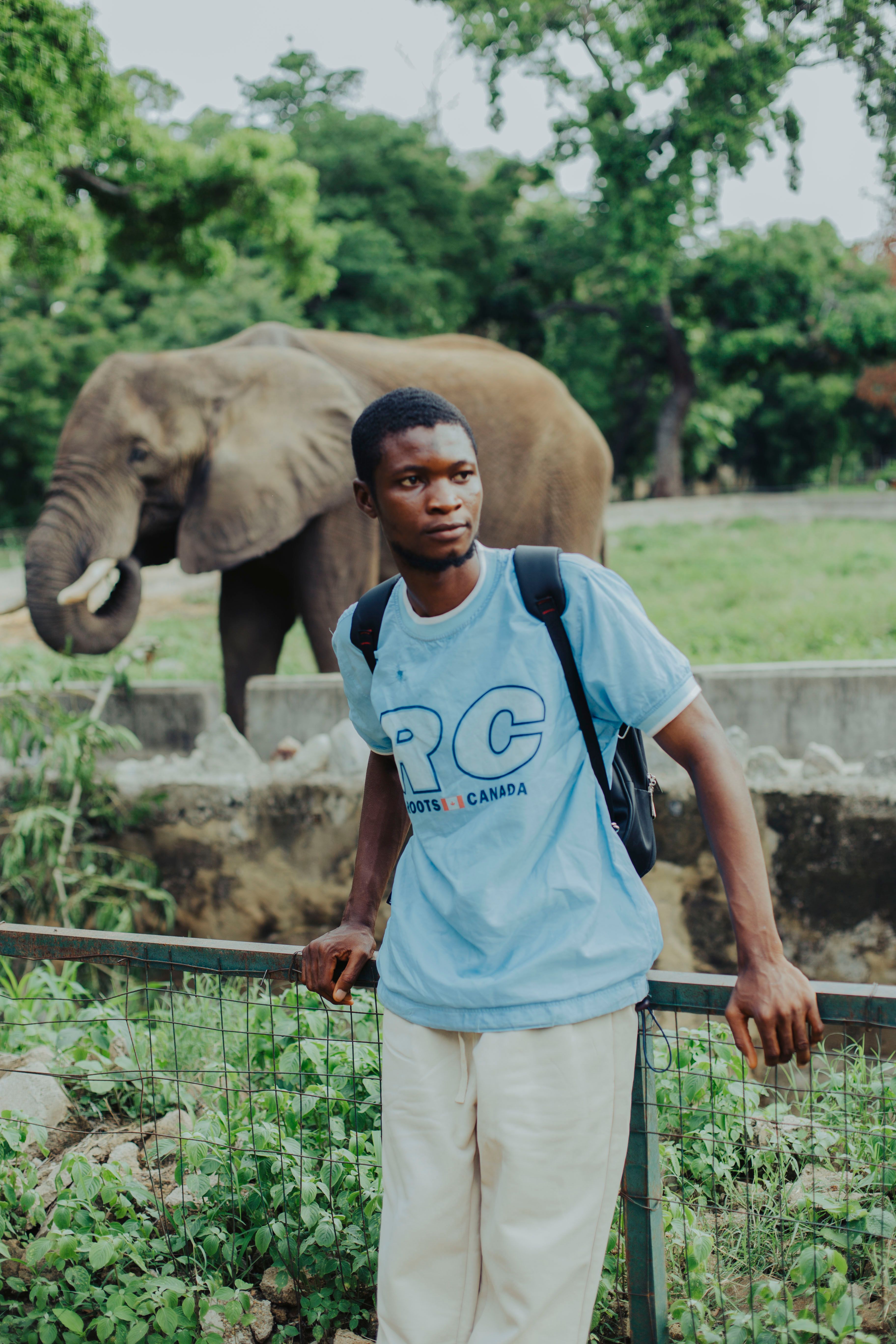 A young man stands by a fence with an elephant in the background at a zoo.