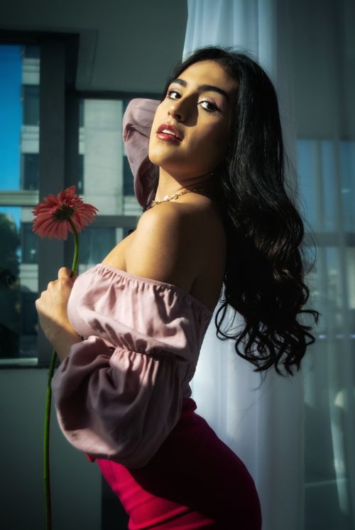 Free Elegant woman in pink gown holding a flower indoors, embracing natural light. Stock Photo