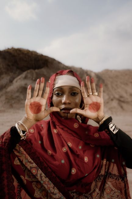 Portrait of a woman in red traditional dress showing hennaed palms in the Nigerian desert setting.