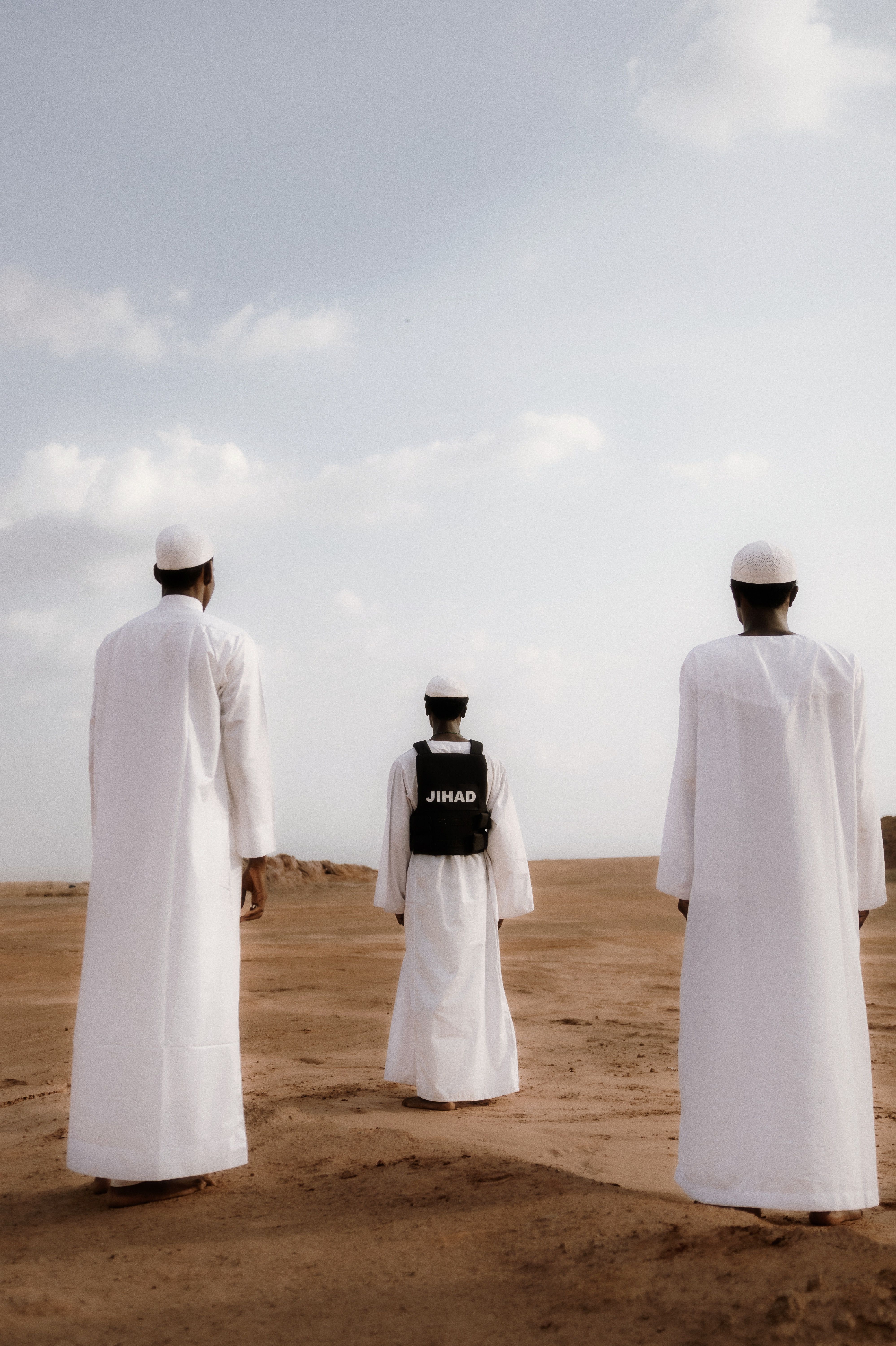 Three men in traditional white garments walking in a desert landscape under a cloudy sky.