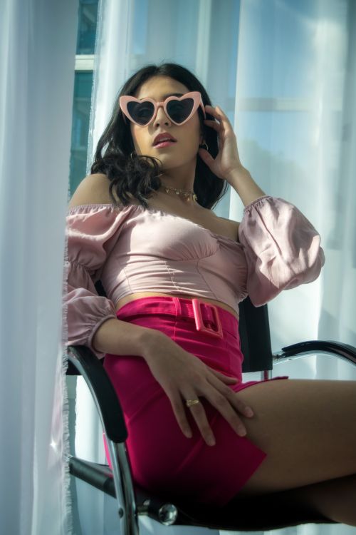 Free Stylish woman posing in heart-shaped sunglasses and chic pink attire indoors. Stock Photo