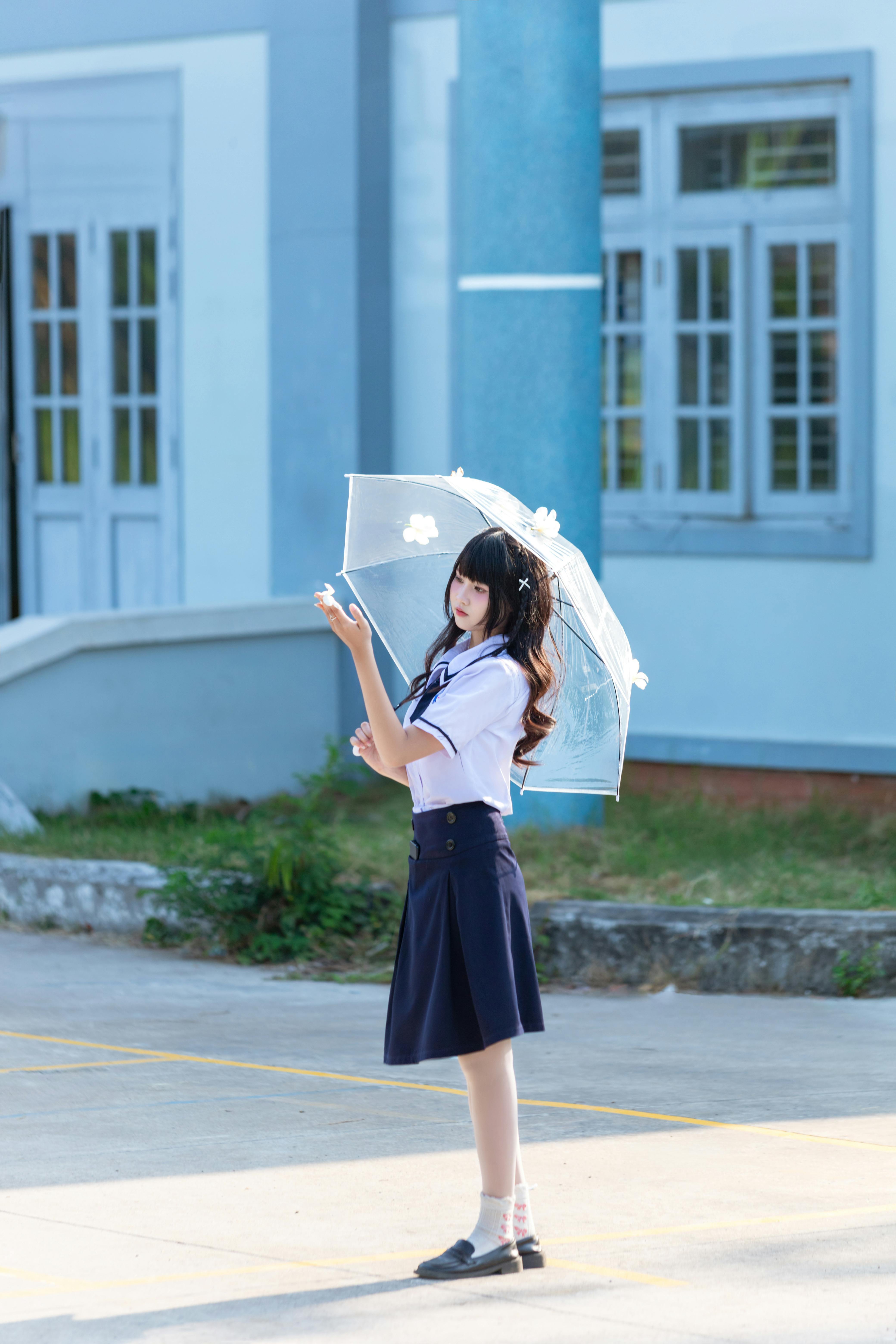 A young woman in school uniform holding a transparent umbrella outdoors, showcasing fashion and casual style.