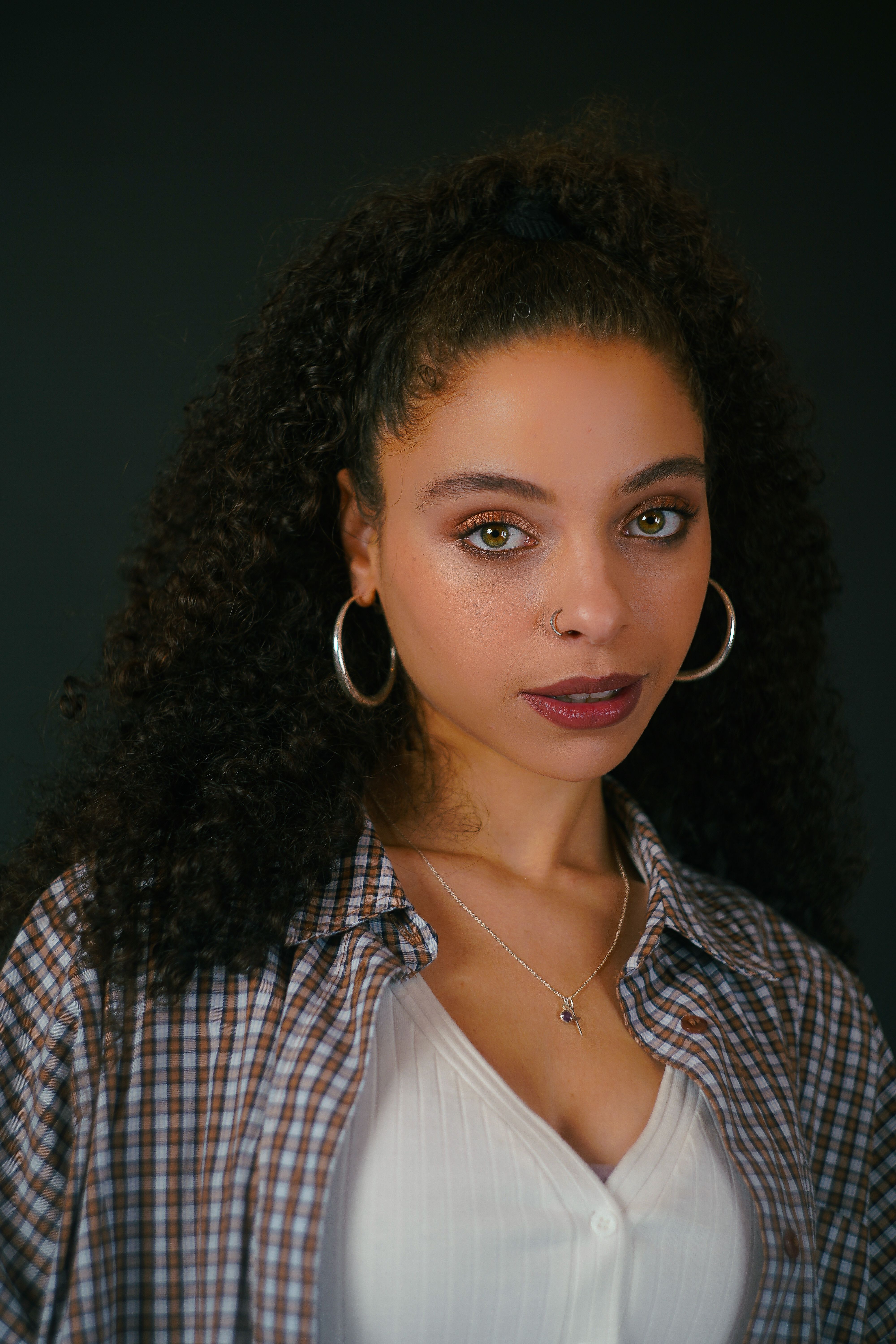 Studio portrait of a woman with curly hair and amber eyes wearing hoop earrings.