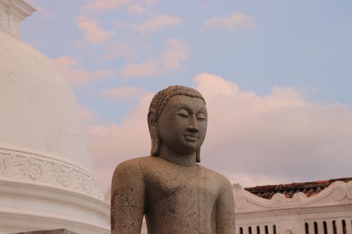 Free A tranquil stone Buddha statue against a vibrant sunrise sky near a white stupa. Stock Photo