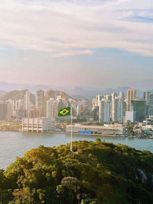 Free Aerial view of Brazilian flag atop green hill with city skyline in the background. Stock Photo