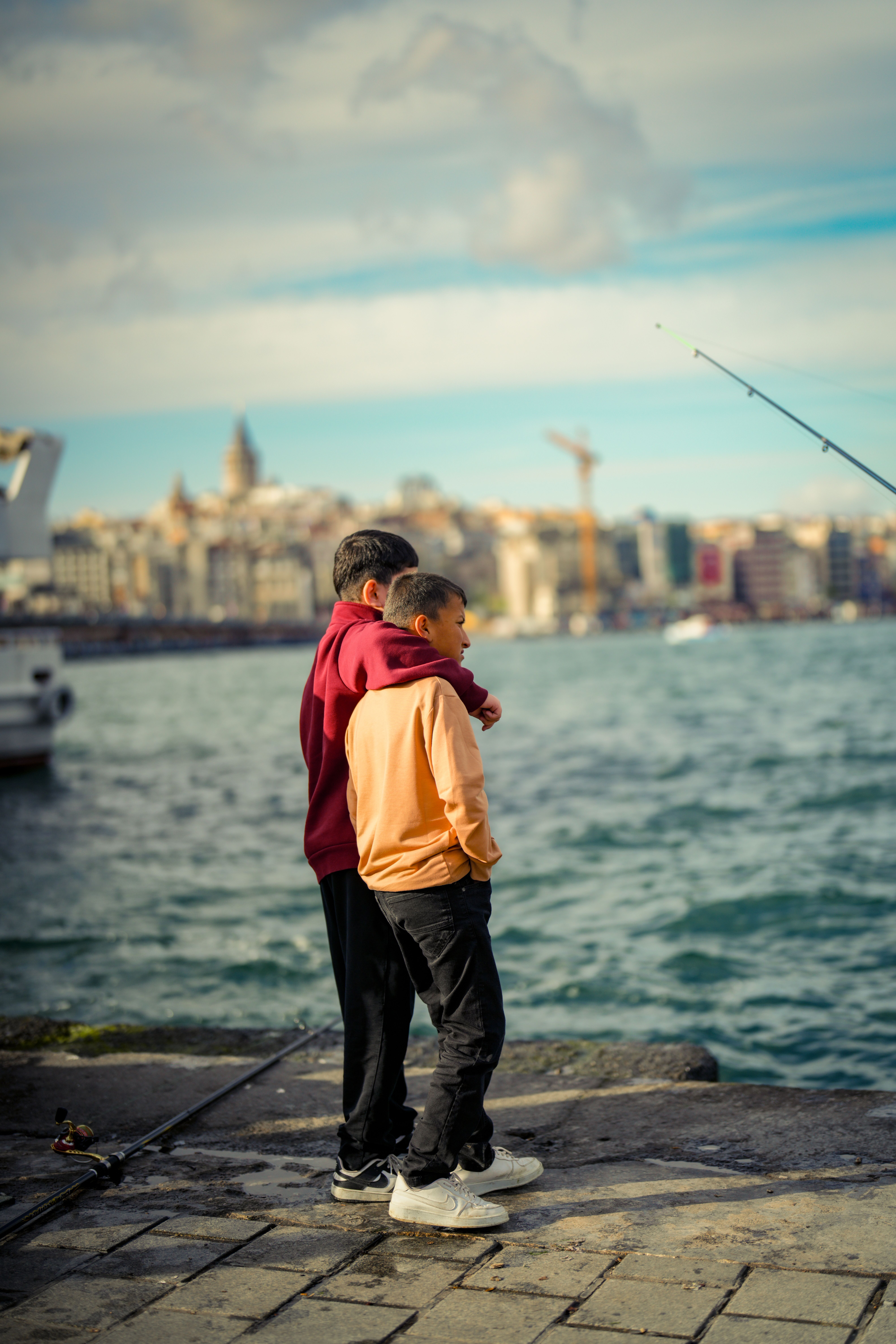 Two kids on a pier looking out over the water in an urban setting, fishing rod nearby.