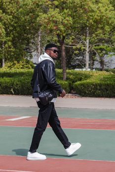 Casually dressed man in urban fashion walking on a sunny basketball court with trees in the background.