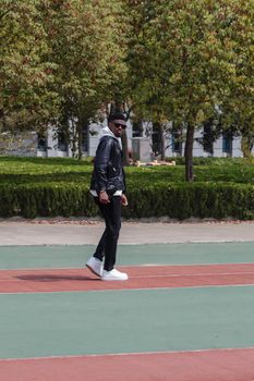 A fashionable man in sunglasses walks across a colorful sports court surrounded by trees.