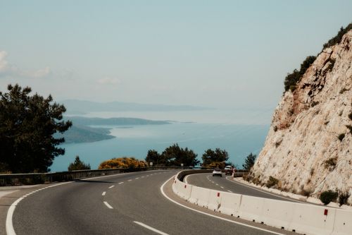 Free Winding road hugging a rocky cliff with stunning ocean view on a sunny day. Stock Photo
