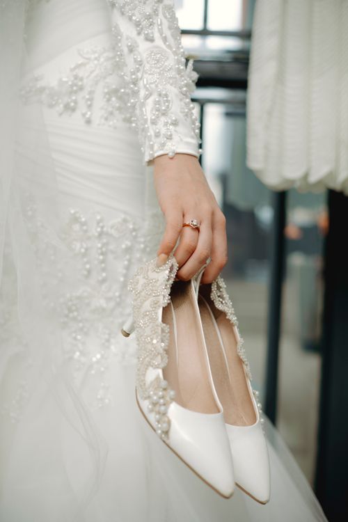 Free Close-up of a bride holding elegant pearl-embellished heels with a lace wedding dress background. Stock Photo