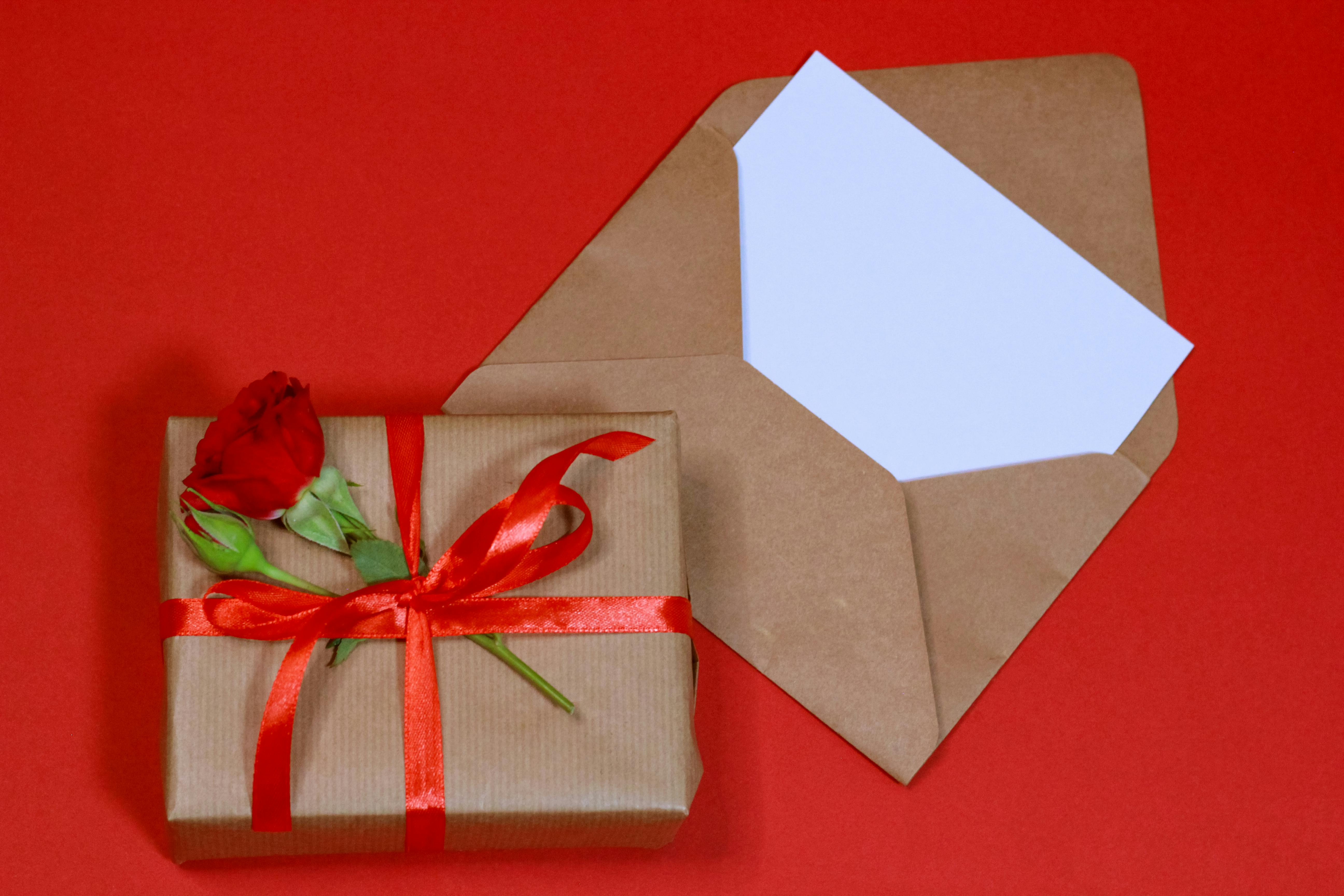 Free A beautifully wrapped gift box with a red rose on a vibrant red background, perfect for celebrations. Stock Photo