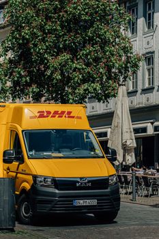 A bright yellow DHL delivery van in a lively European town square, showcasing a mix of urban and traditional architecture.