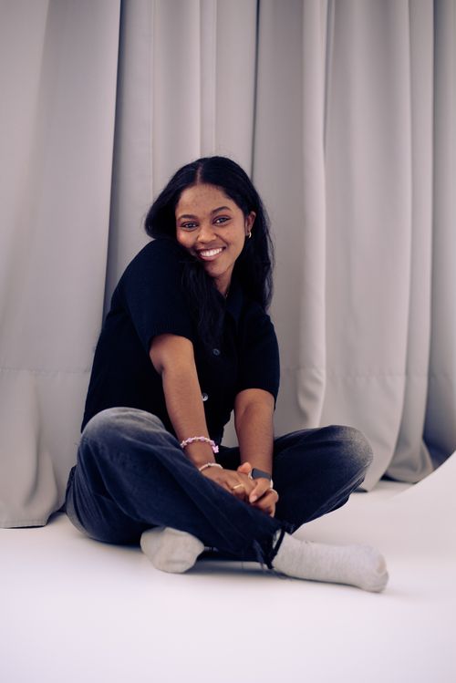 Free Smiling young woman in casual attire sitting indoors against a curtain backdrop, exuding joy and relaxation. Stock Photo