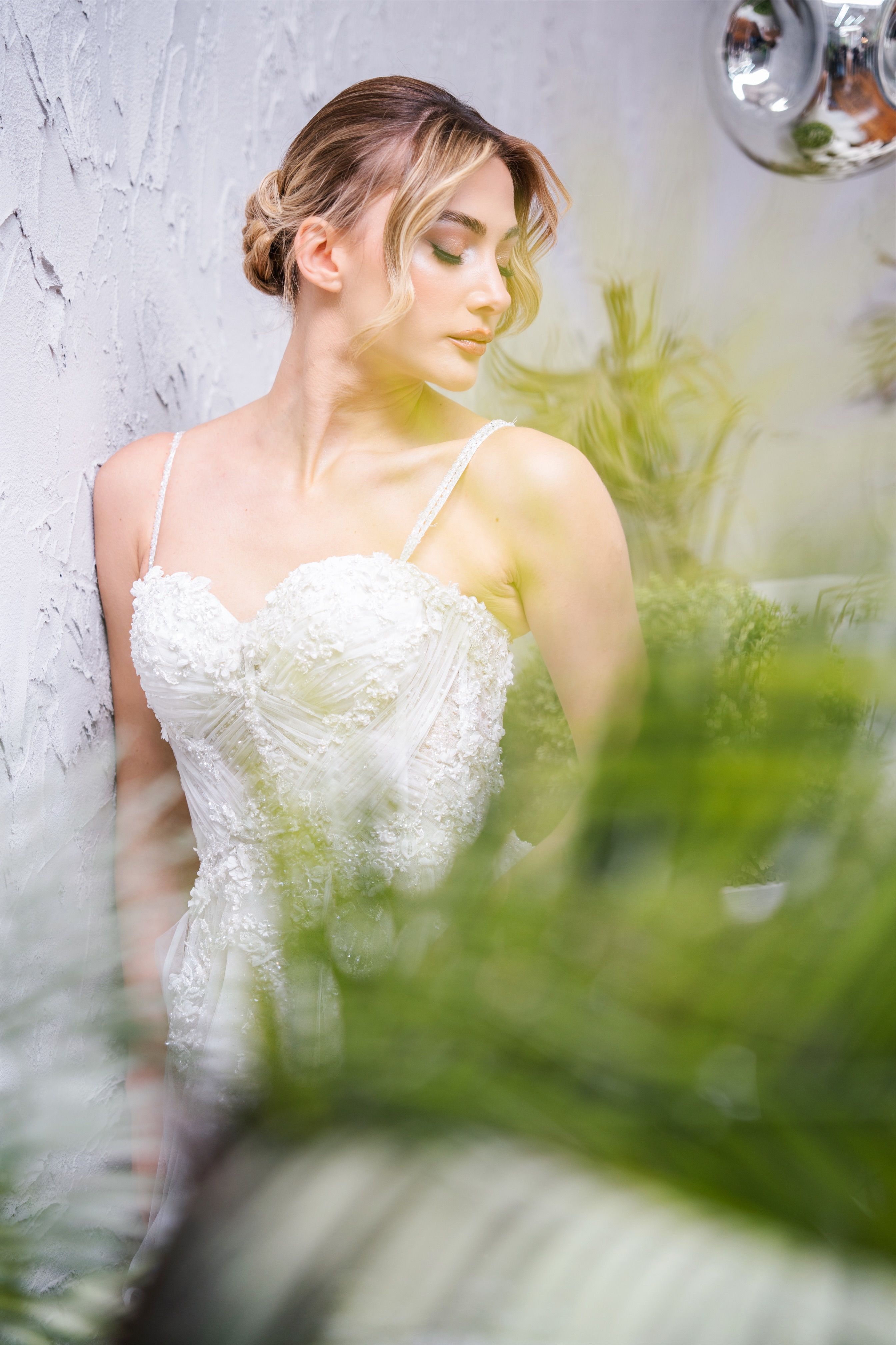 Stylish bride in a white gown against a textured wall surrounded by greenery.