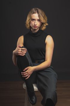 Portrait of a stylish woman sitting on a stool in a black sleeveless top against a dark background.