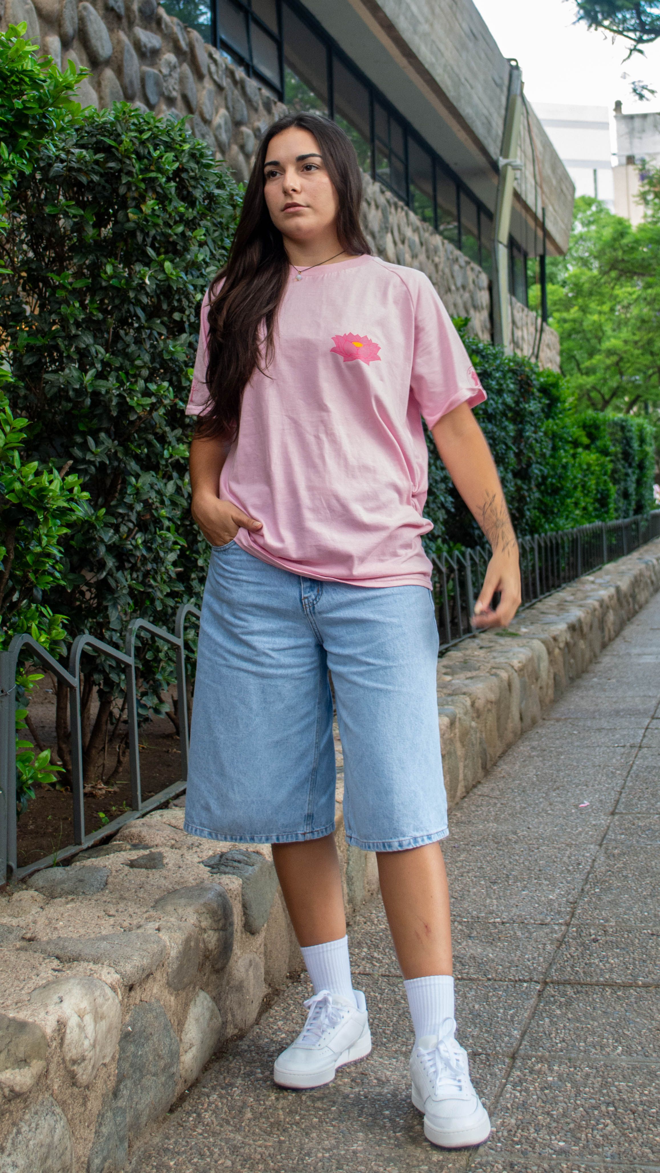 Young adult in casual streetwear, featuring pink tee and denim shorts on city sidewalk.
