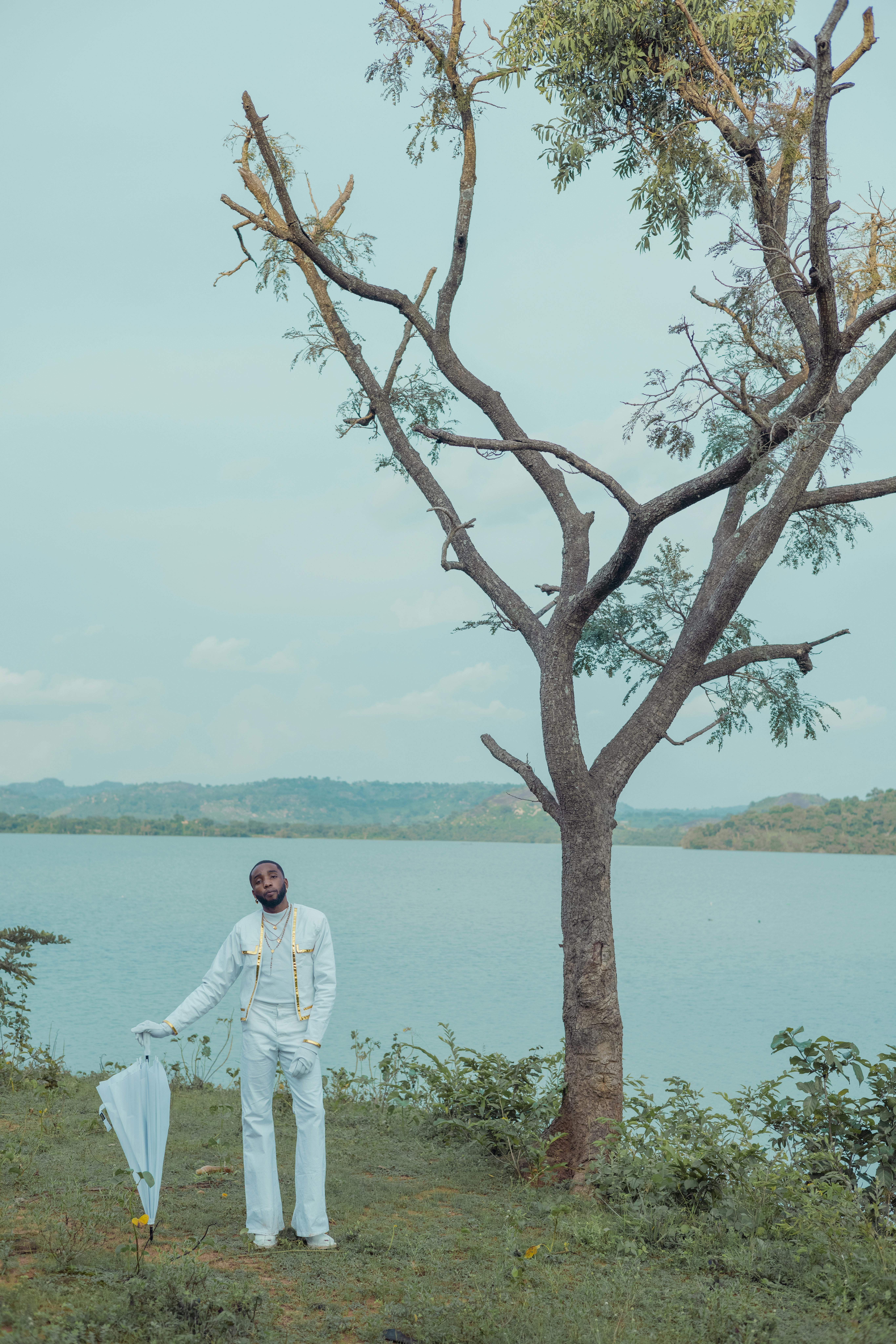 A fashionable man in formal white stands by a tranquil lakeside with a tree and umbrella.