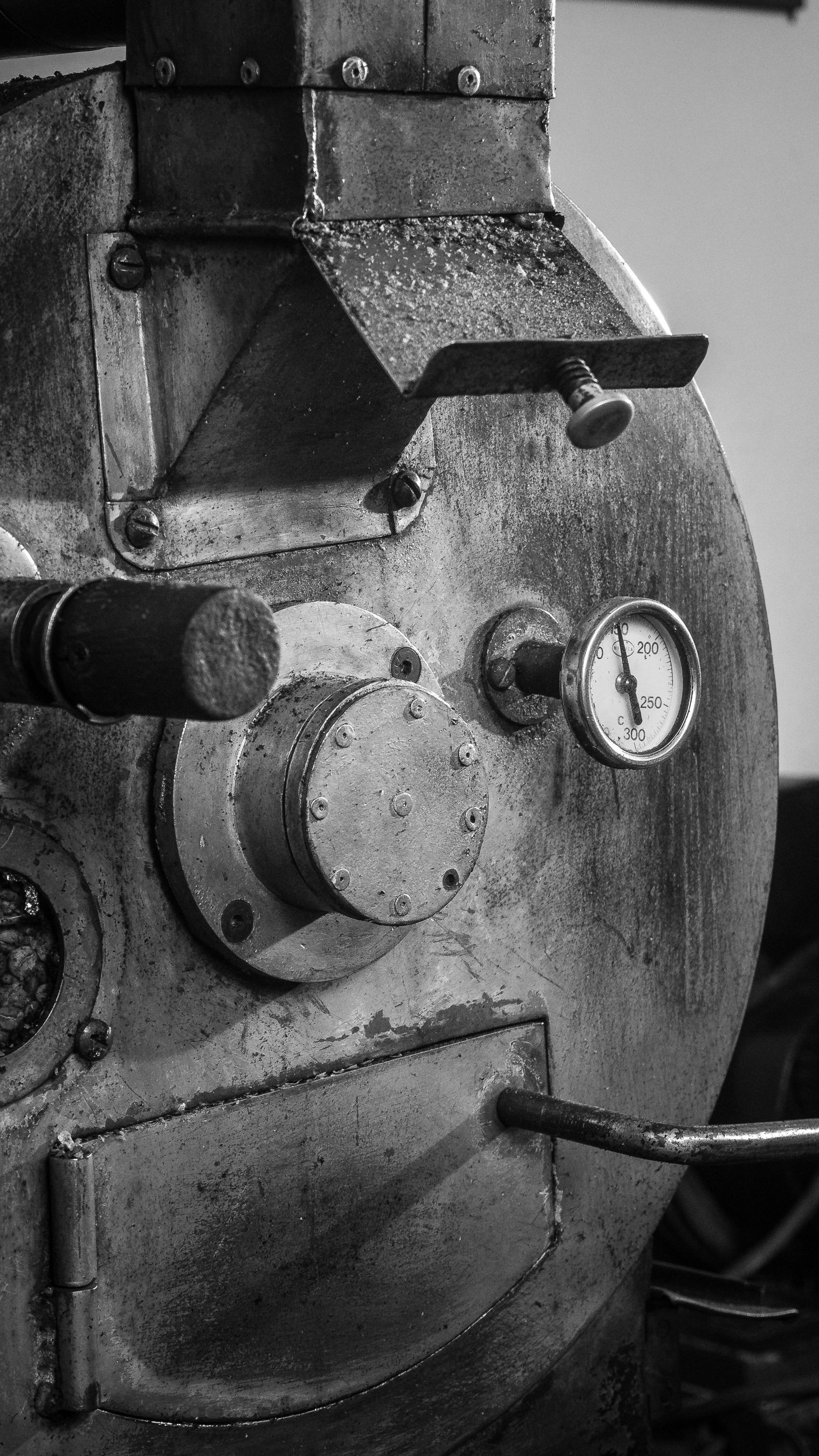 A detailed black and white image of an industrial coffee roaster machine, highlighting gauge and metal textures.