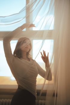 A tranquil scene of a woman gracefully posing by a window with sheer curtains in warm sunlight.