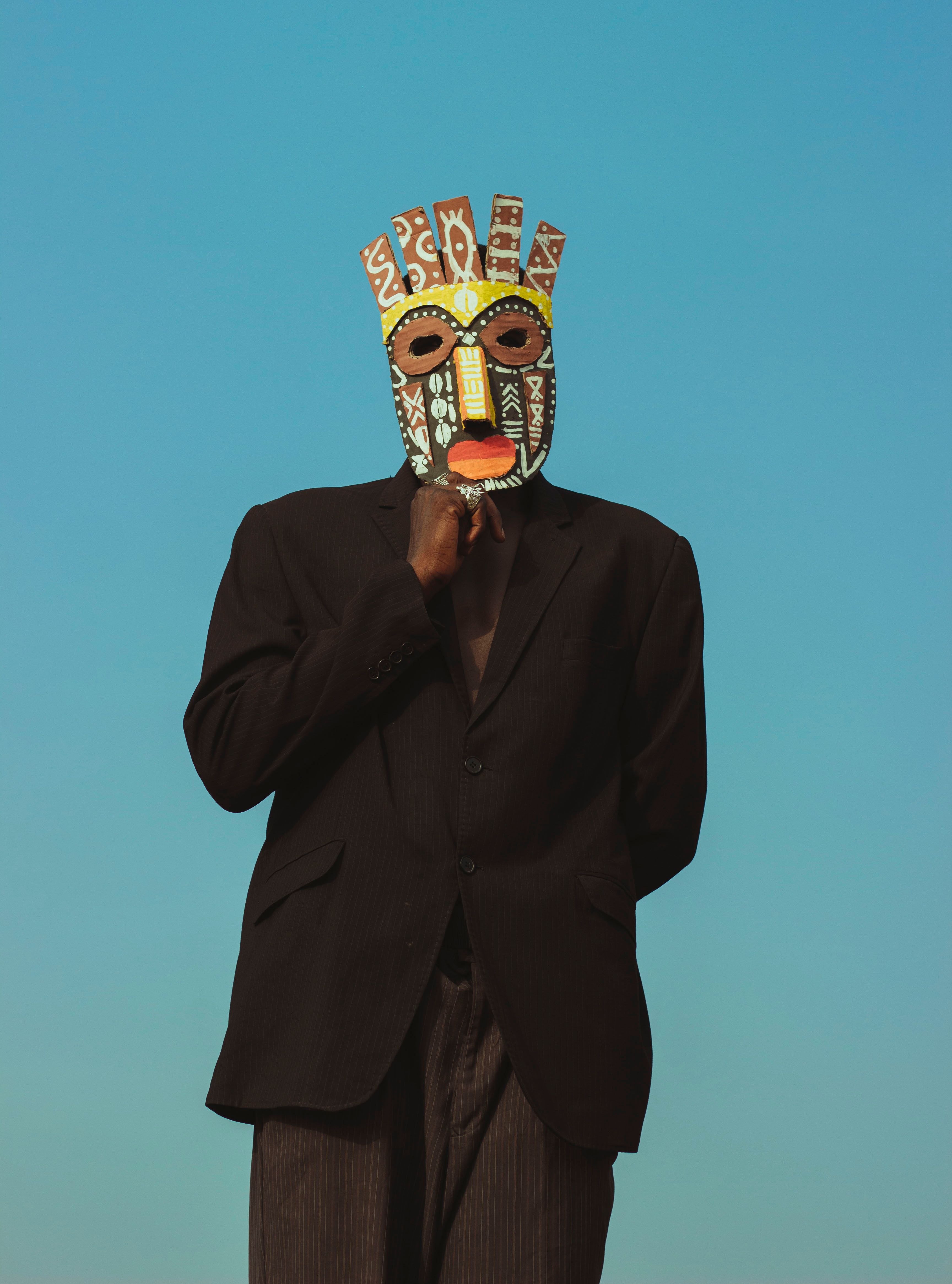 A person in a colorful tribal mask poses against a bright blue sky, conveying cultural and artistic themes.