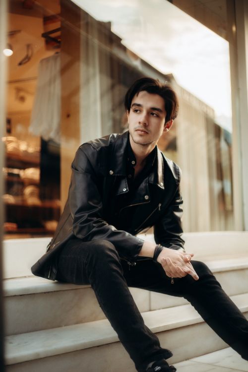 Free Stylish young man in black leather jacket sitting thoughtfully on marble steps outdoors. Stock Photo
