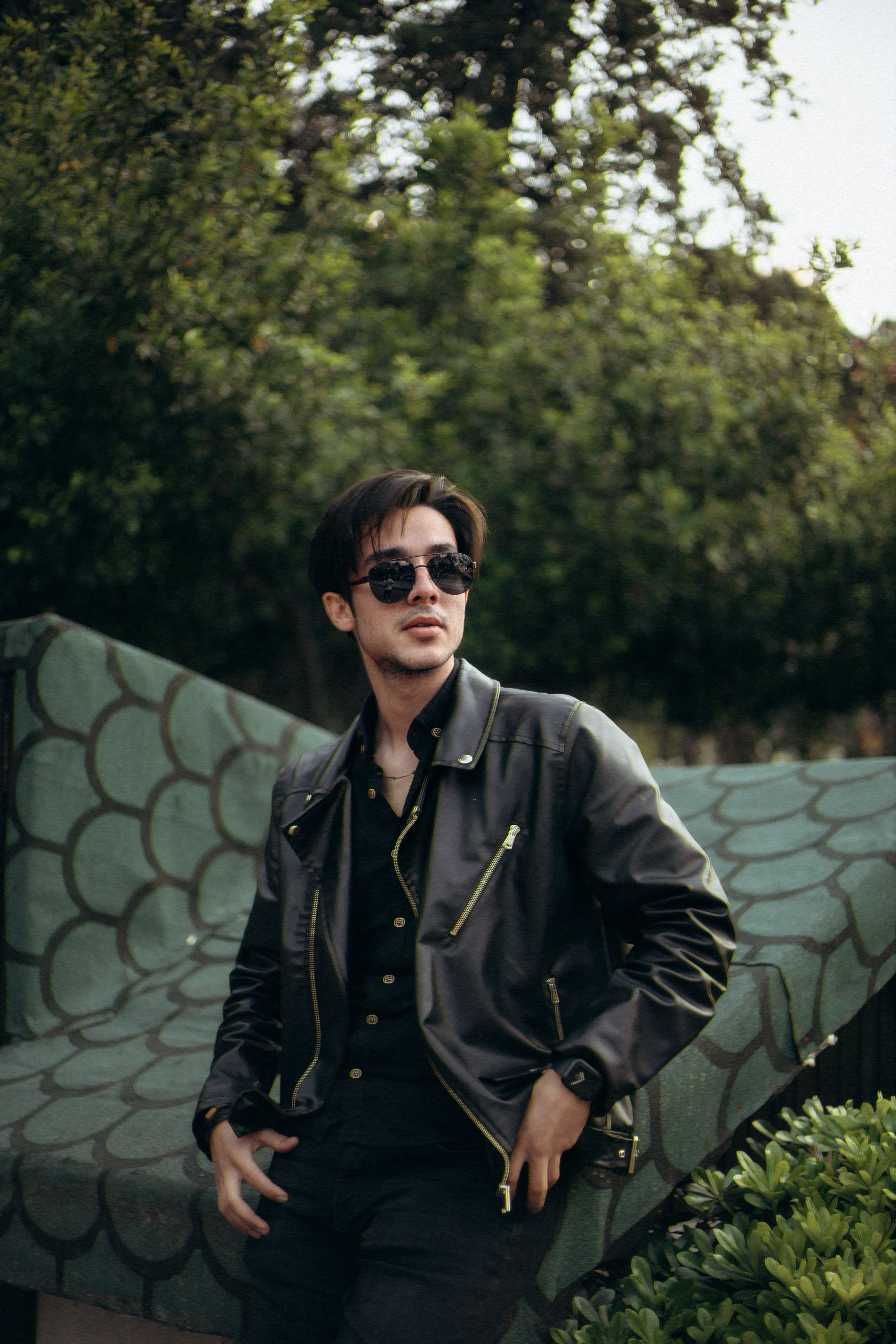 Young man in sunglasses and leather jacket posing confidently outdoors.