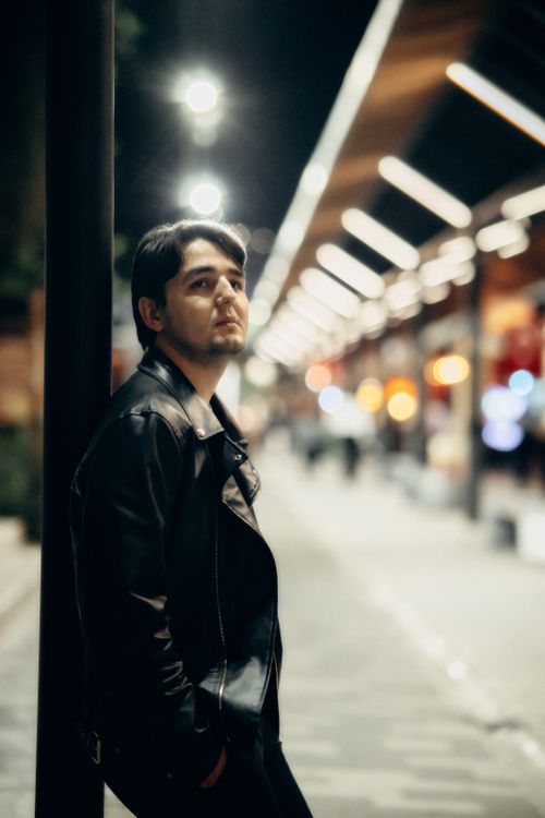 Free Young man in leather jacket leans against pole in a night urban setting. Stock Photo