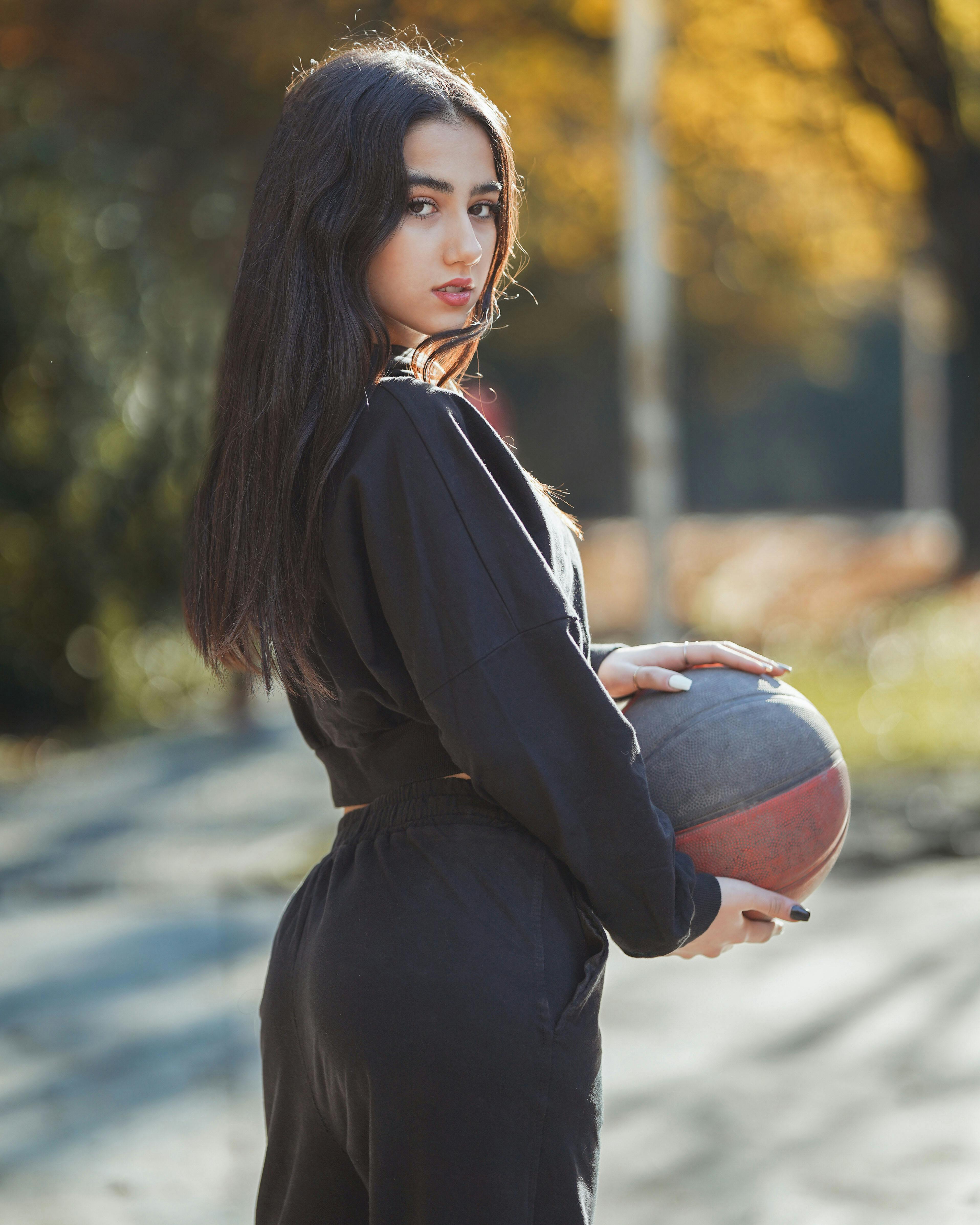 在阳光下，一位时尚的年轻女子自信地拿着篮球，在户外摆姿势