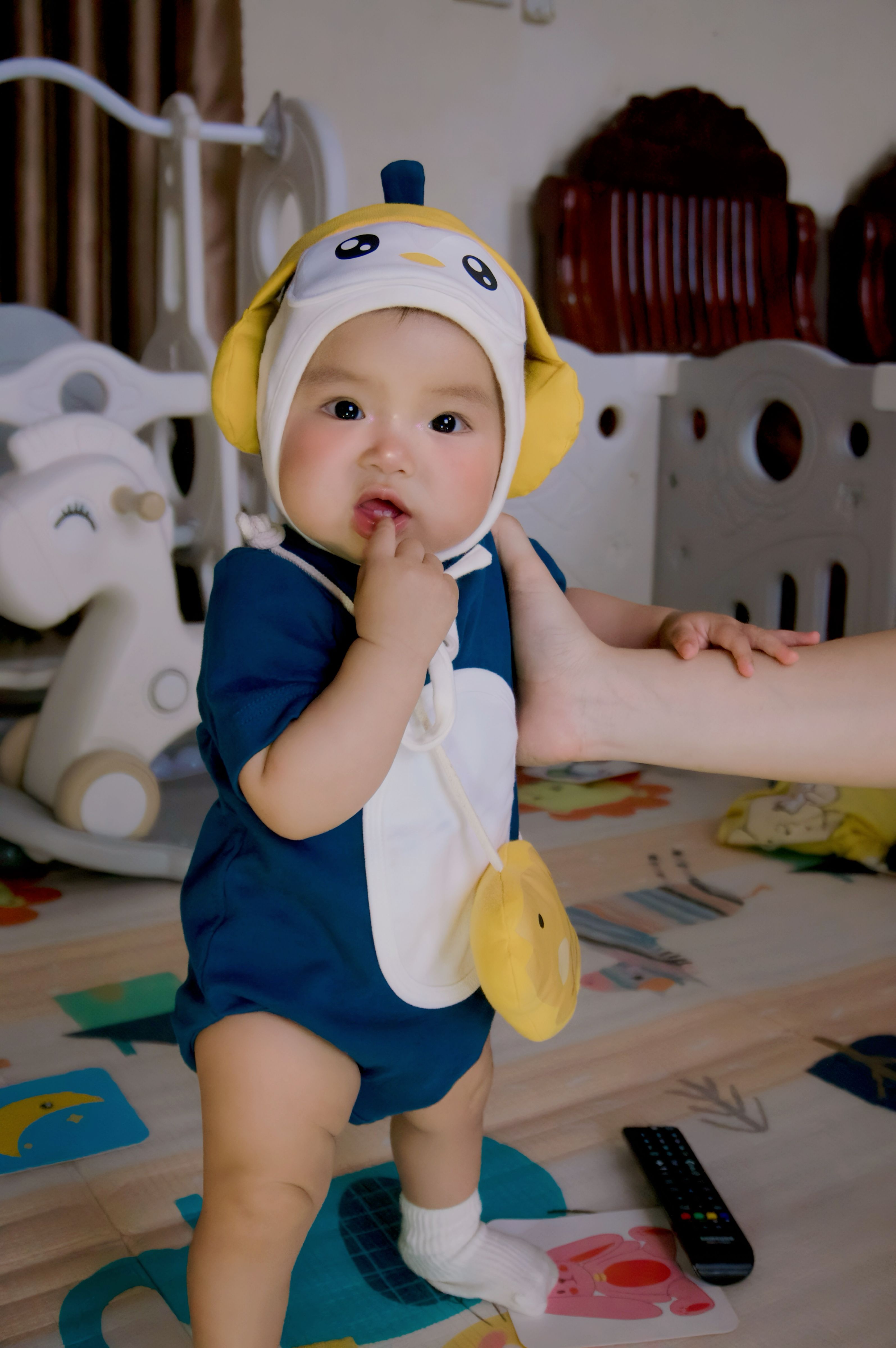 Adorable baby in a penguin outfit, playfully chewing finger indoors. Warm, cozy, and delightful scene.