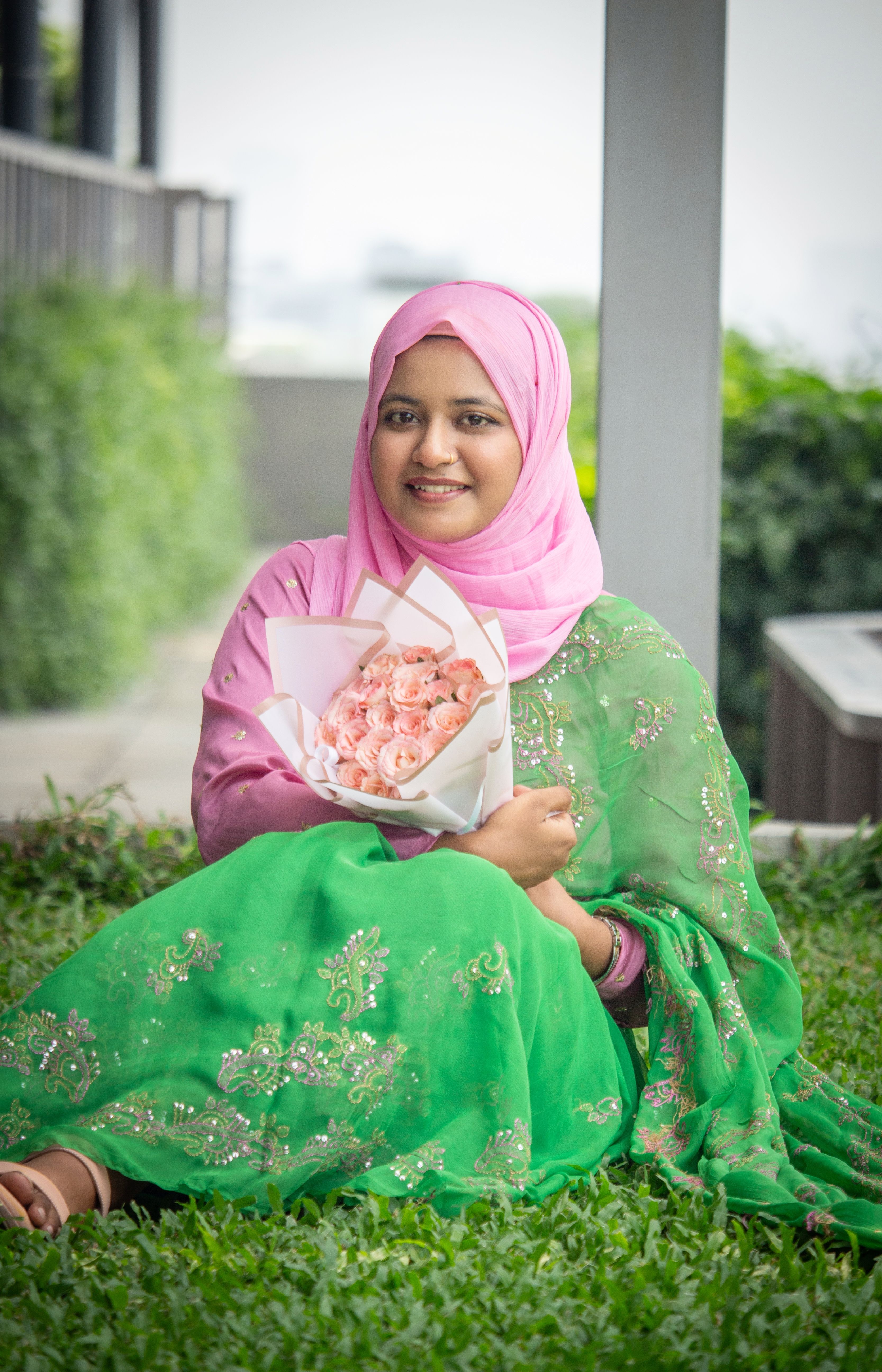 A cheerful woman in a pink hijab holds a rose bouquet while sitting outdoors in a green saree.