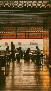 Silhouetted group dining at a rooftop restaurant during a warm sunset overlooking the sea.