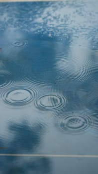 Blue surface with raindrops forming ripples and reflections of clouds.