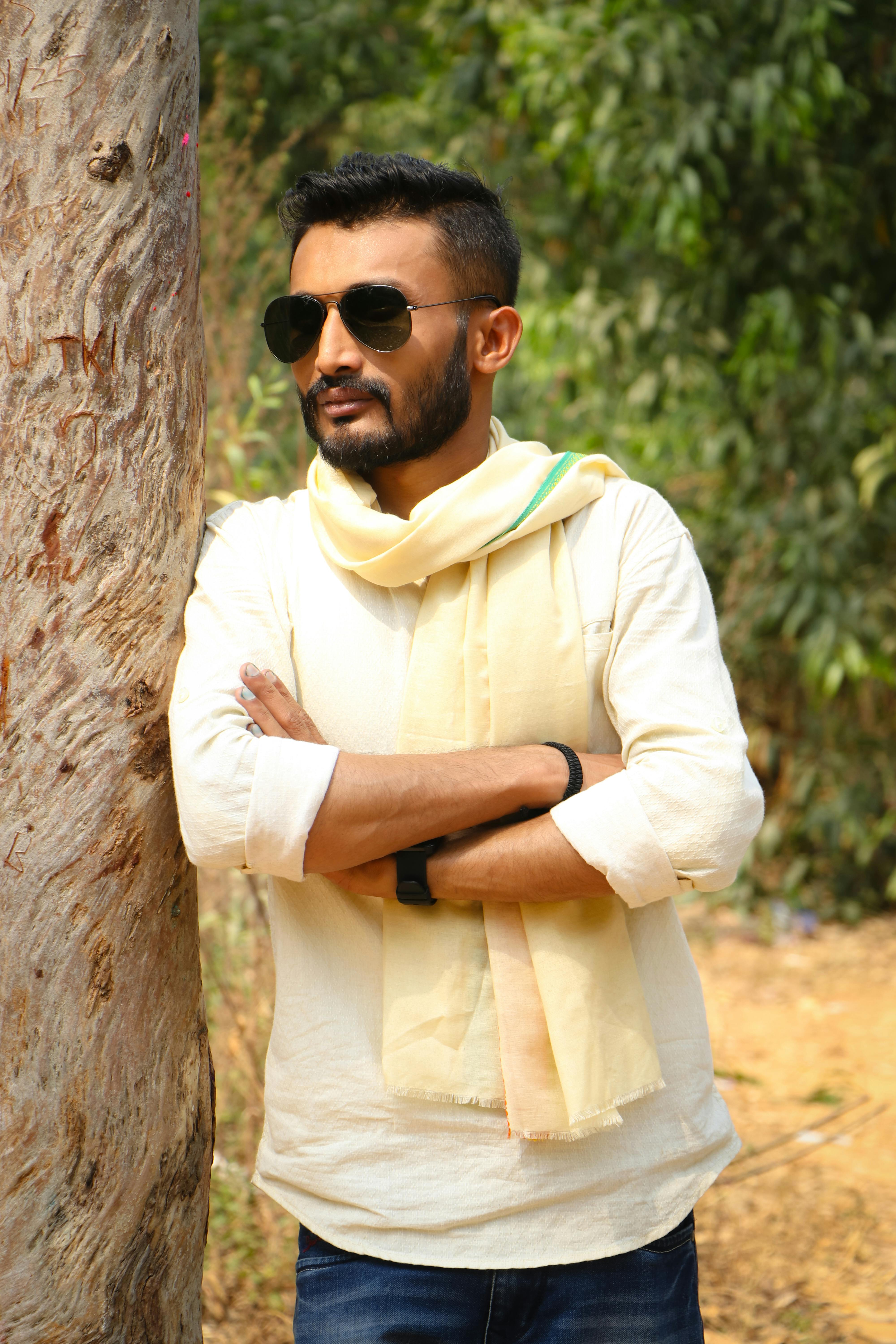 A confident man with sunglasses leaning against a tree in a sunny outdoor setting.