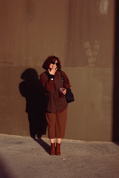 Woman in stylish brown outfit standing and smoking against a sunlit wall.