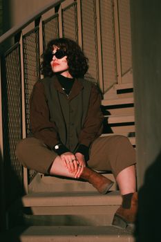 Fashionable woman in sunglasses posing on metal stairs in moody lighting.