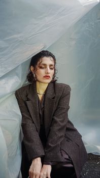 A contemplative woman in a dark coat sits against a plastic backdrop in an artistic indoor setting.