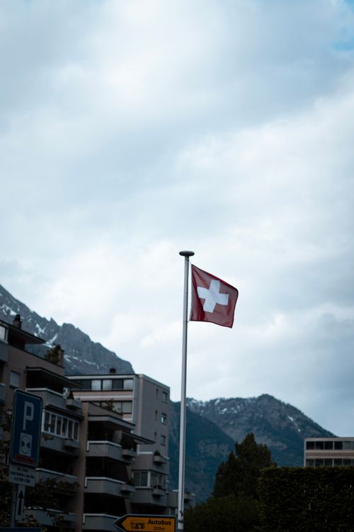 Free Swiss flag flying in a town with mountains and cloudy sky in background. Stock Photo