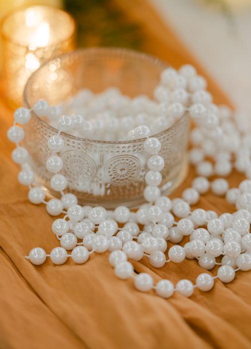 Free A glass bowl filled with white beaded garland, lit by warm candlelight on a fabric backdrop. Stock Photo