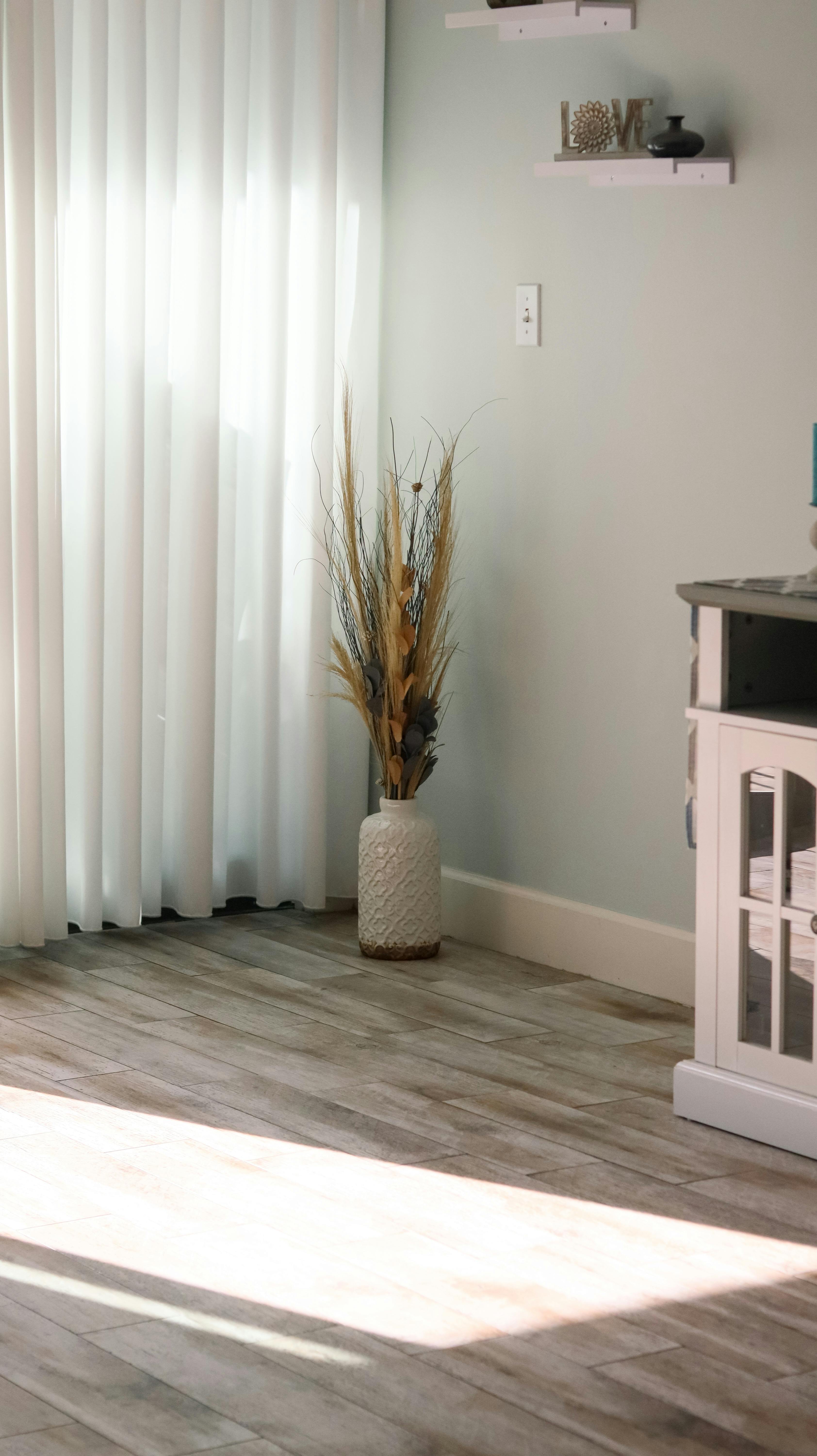 Sunlit corner of a minimalist living room featuring a decorative vase, elegant curtains, and stylish flooring.