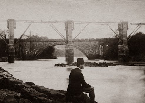 这是一张复古风格的黑白照片，照片中一名男子站在河边，身旁是一座历史悠久的桥梁