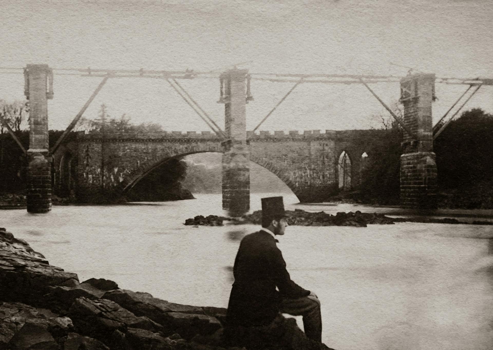 Grayscale Photo Man Wearing A Top Hat And Suit Sitting By The River Near A Bridge