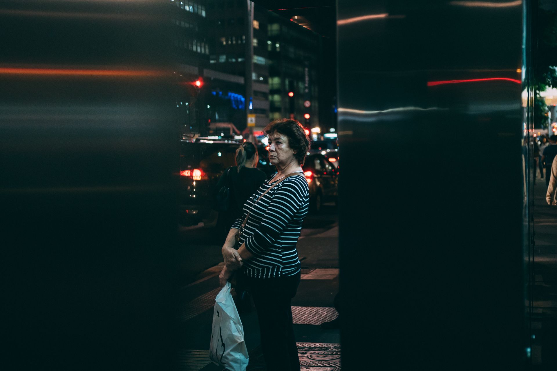 An older woman stands on a bustling street in São Paulo at night, holding a bag.
