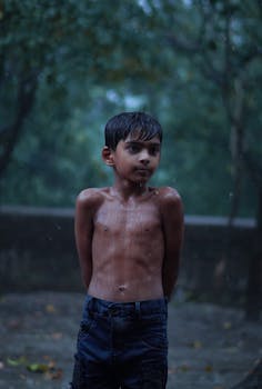 Wet shirtless boy standing in rain with a serene expression, surrounded by lush greenery.