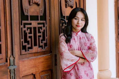 Free A woman in a pink floral qipao stands by intricately carved wooden doors. Stock Photo